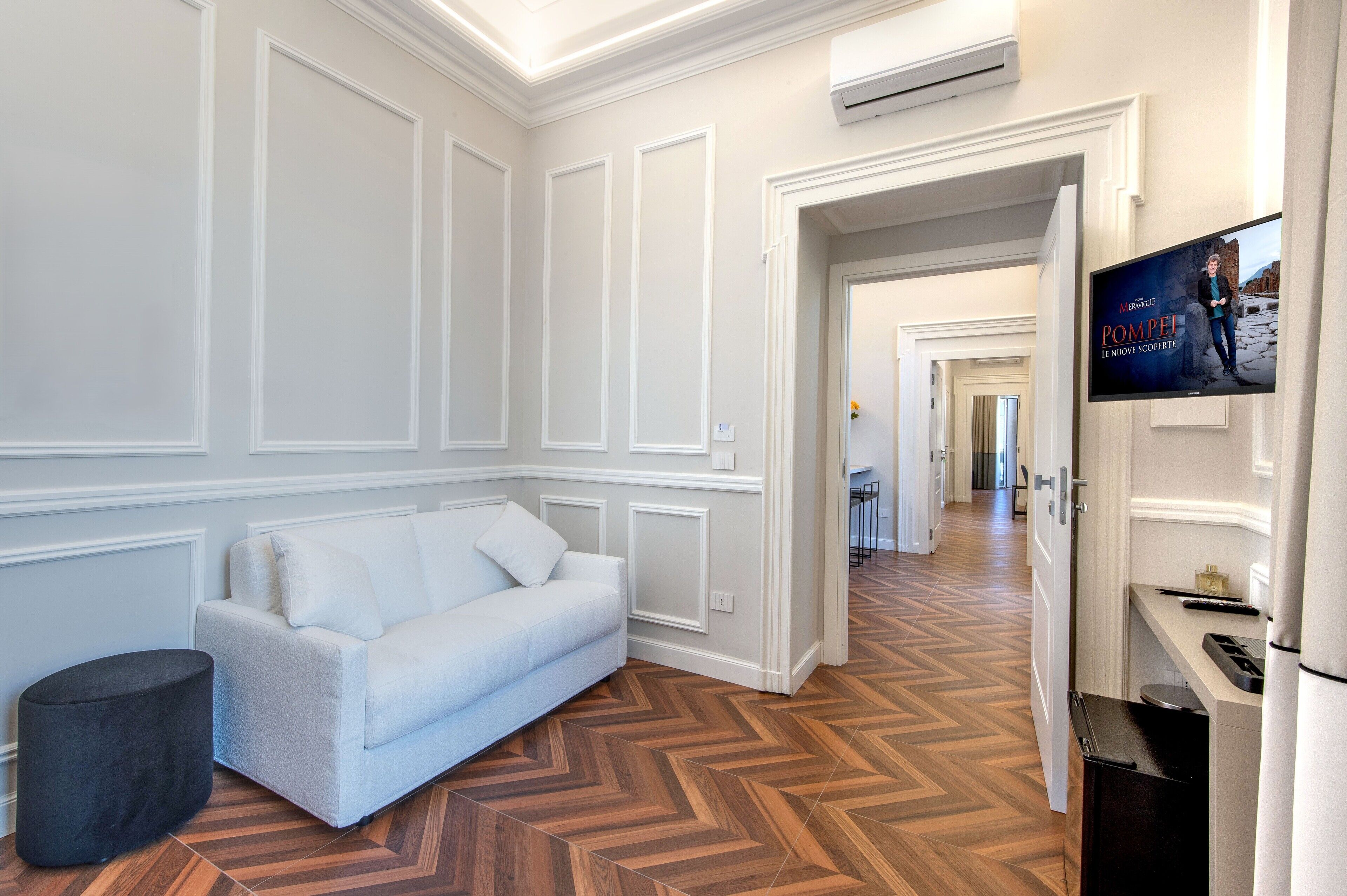 Ein Hotelzimmer mit einem weißen Sofa, einem schwarzen Hocker, einem Holzboden mit Fischgrätmuster und einem an der Wand befestigten Fernseher.