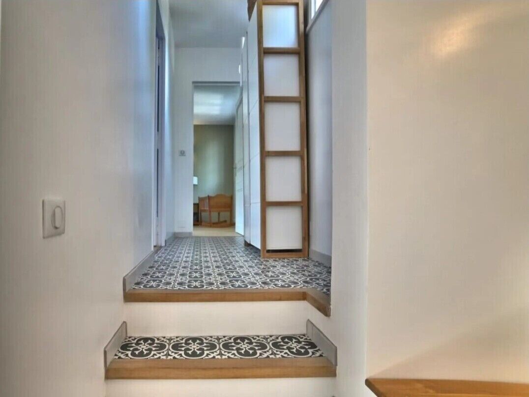 Un couloir étroit, dont le sol est recouvert de carreaux, mène à un escalier en bois et à une échelle.