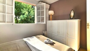 Un dormitorio con cama, ventana con vista a árboles, armario y lámpara.