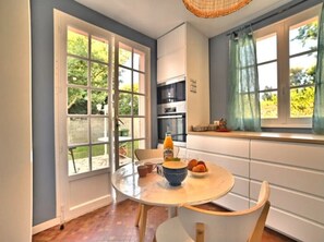 Una cocina con una mesa redonda, un tazón de fruta y una botella de jugo de naranja. Hay dos ventanas con cortinas, y se ve follaje al exterior.