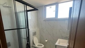 Un baño con cabina de ducha de vidrio, inodoro, lavamanos y ventana.