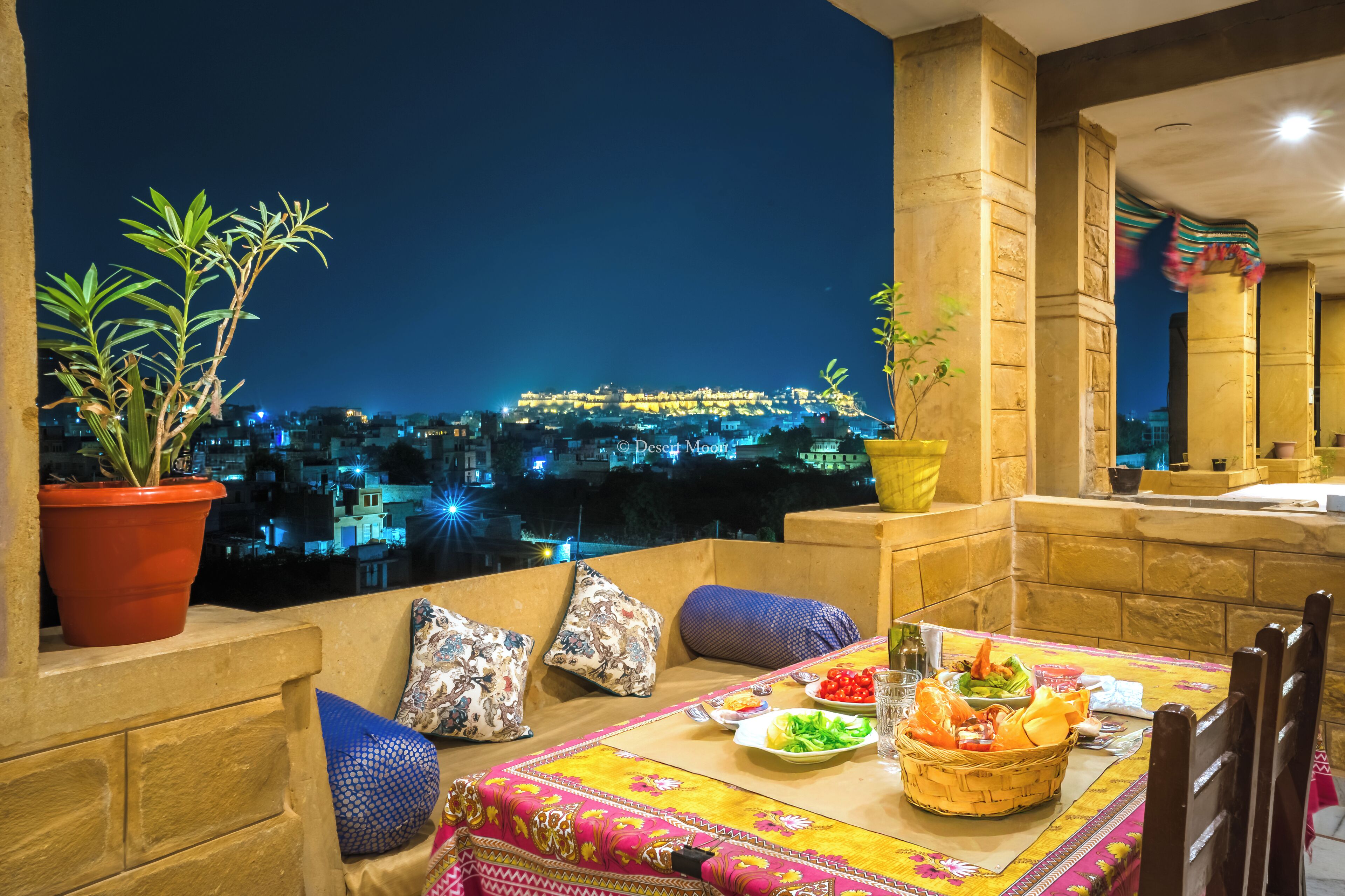 A balcony with a table set for dining, potted plants, and a view of a cityscape at night.