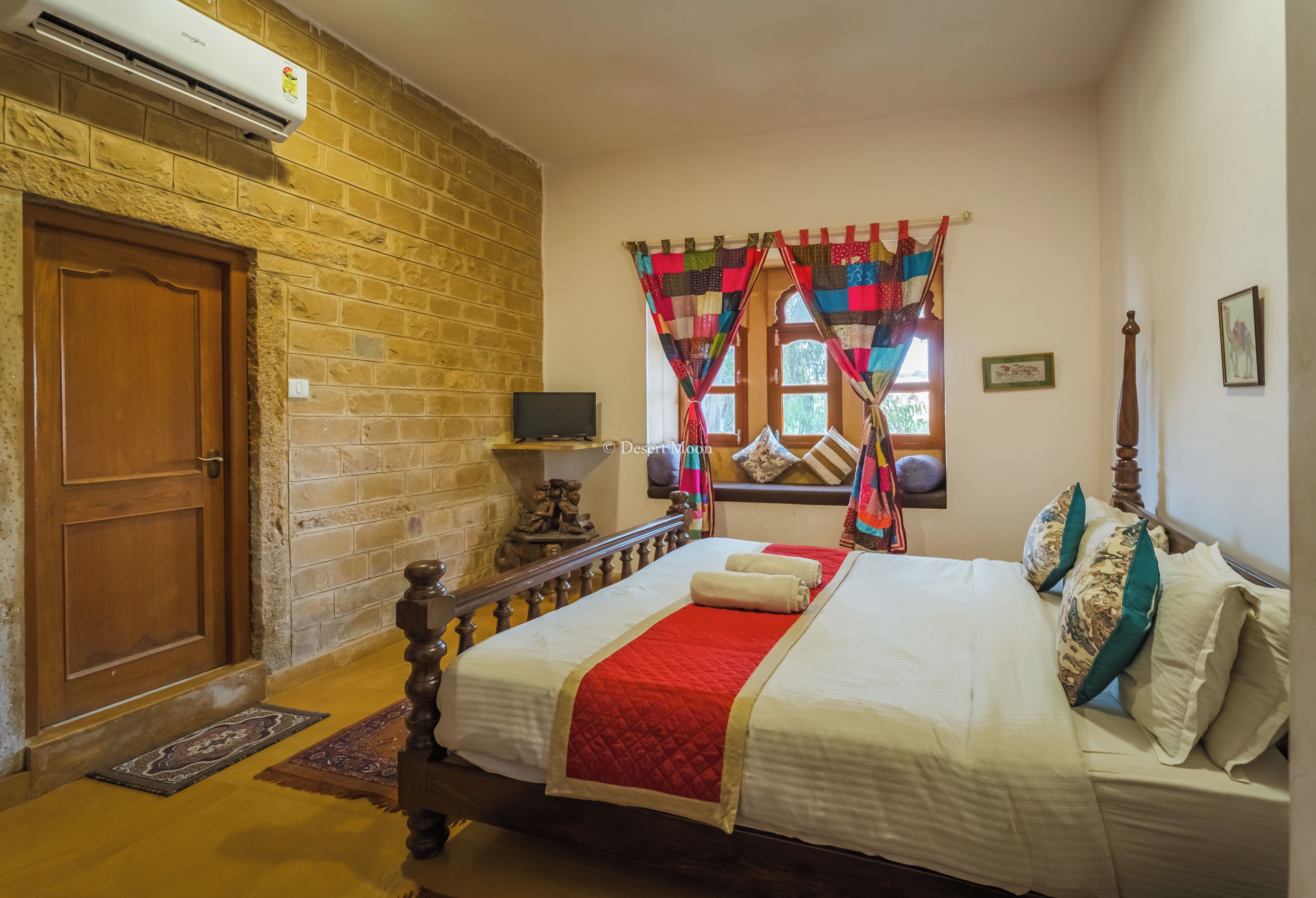 A bedroom with a brick wall, a wooden door, a bed with a red blanket, a TV, and a window with colorful curtains.