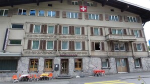 Un edificio de varios pisos con una bandera suiza, un letrero de restaurante y mesas con sillas en su terraza.