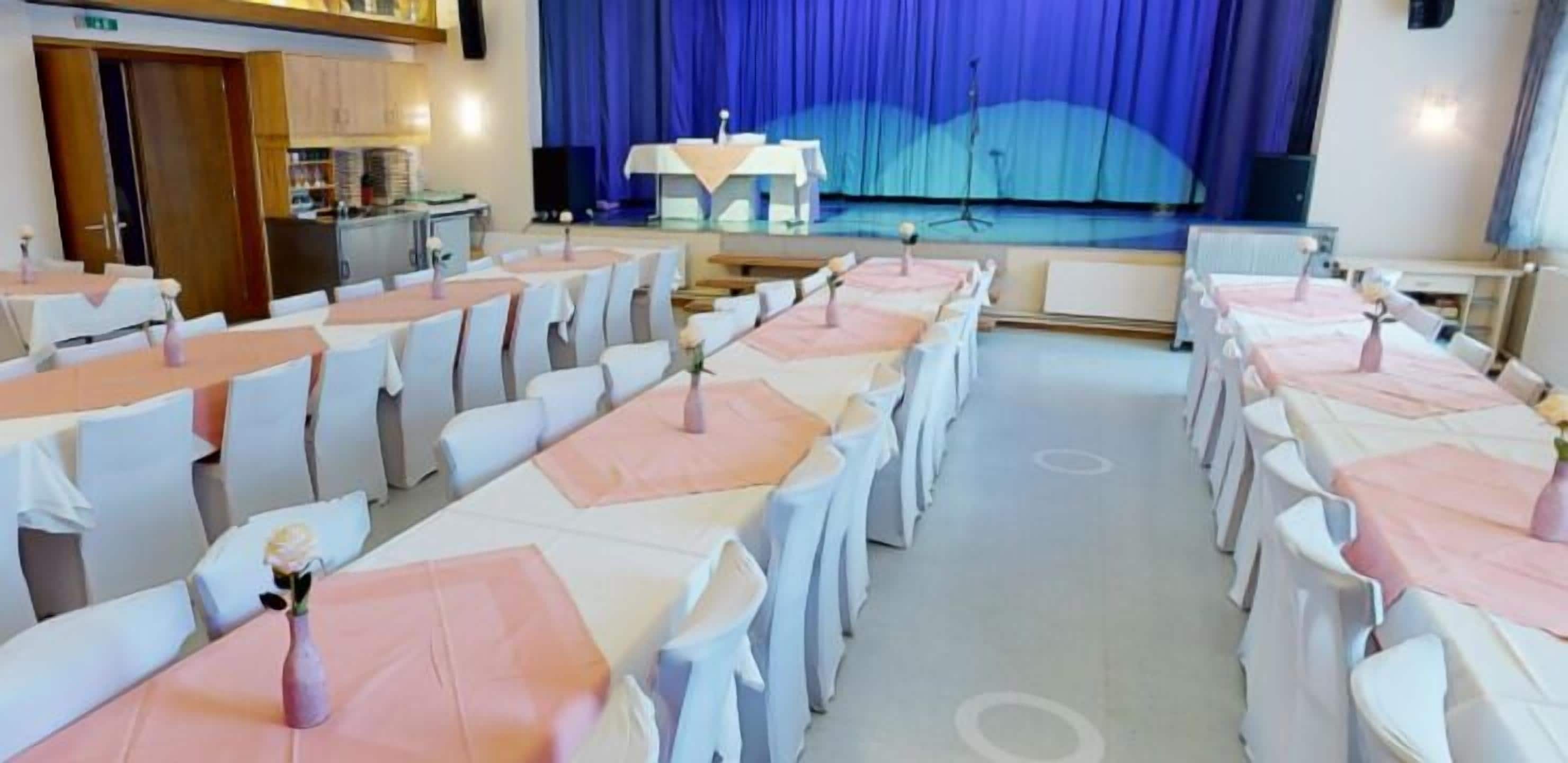 A conference room with rows of tables and chairs, a stage with a blue curtain backdrop, and a kitchenette area.