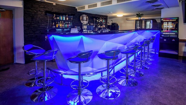 Una zona de bar con un mostrador iluminado en azul, varios taburetes y una máquina tragamonedas al fondo.