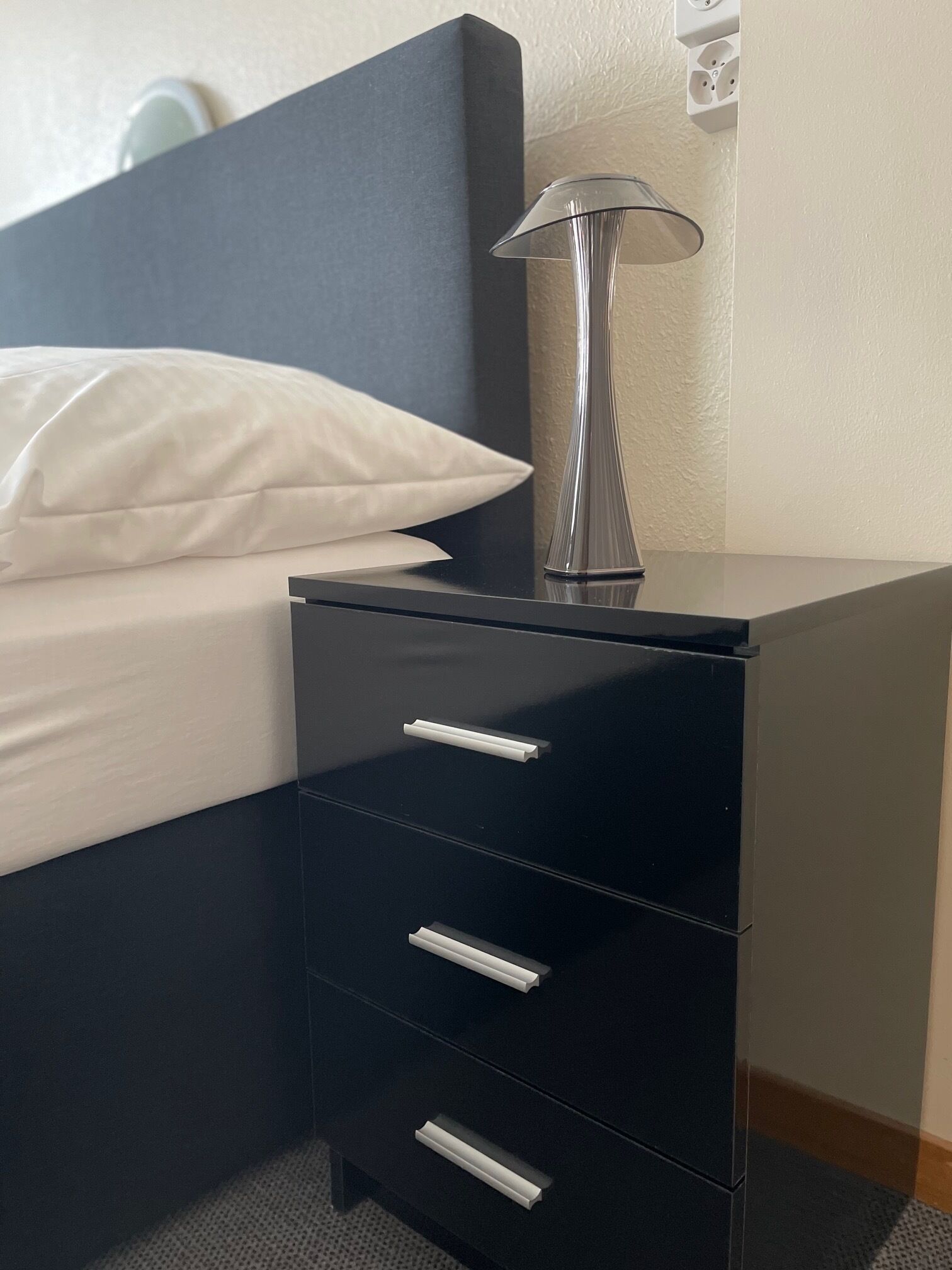 Une table de chevet noire avec une lampe, un oreiller blanc et un lit.
