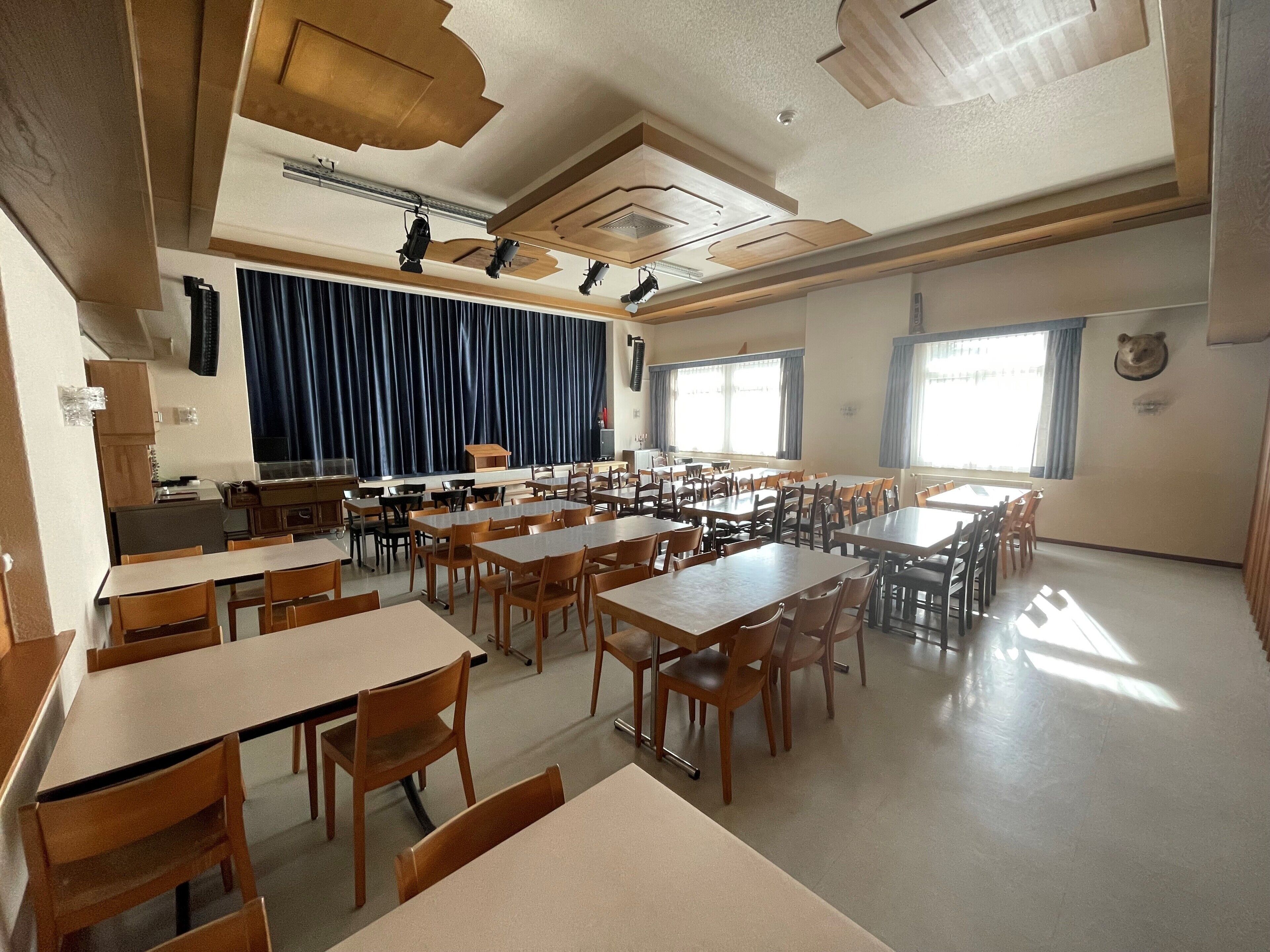A classroom with multiple wooden tables and chairs, a stage with curtains, and a bear head decoration on the wall.