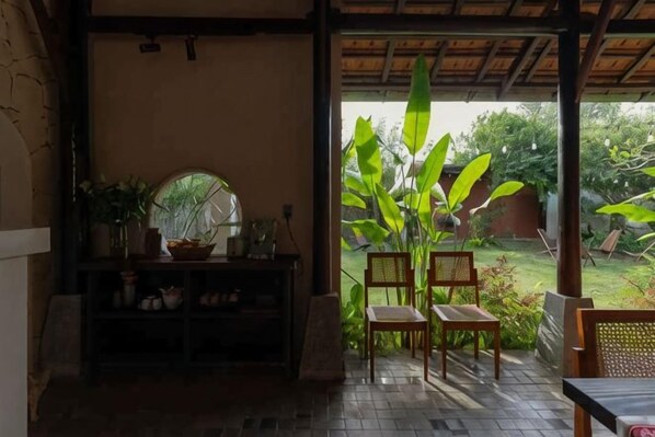 A room with a wooden bench, a table, a shelf with various items, and a view of a garden with large plants.