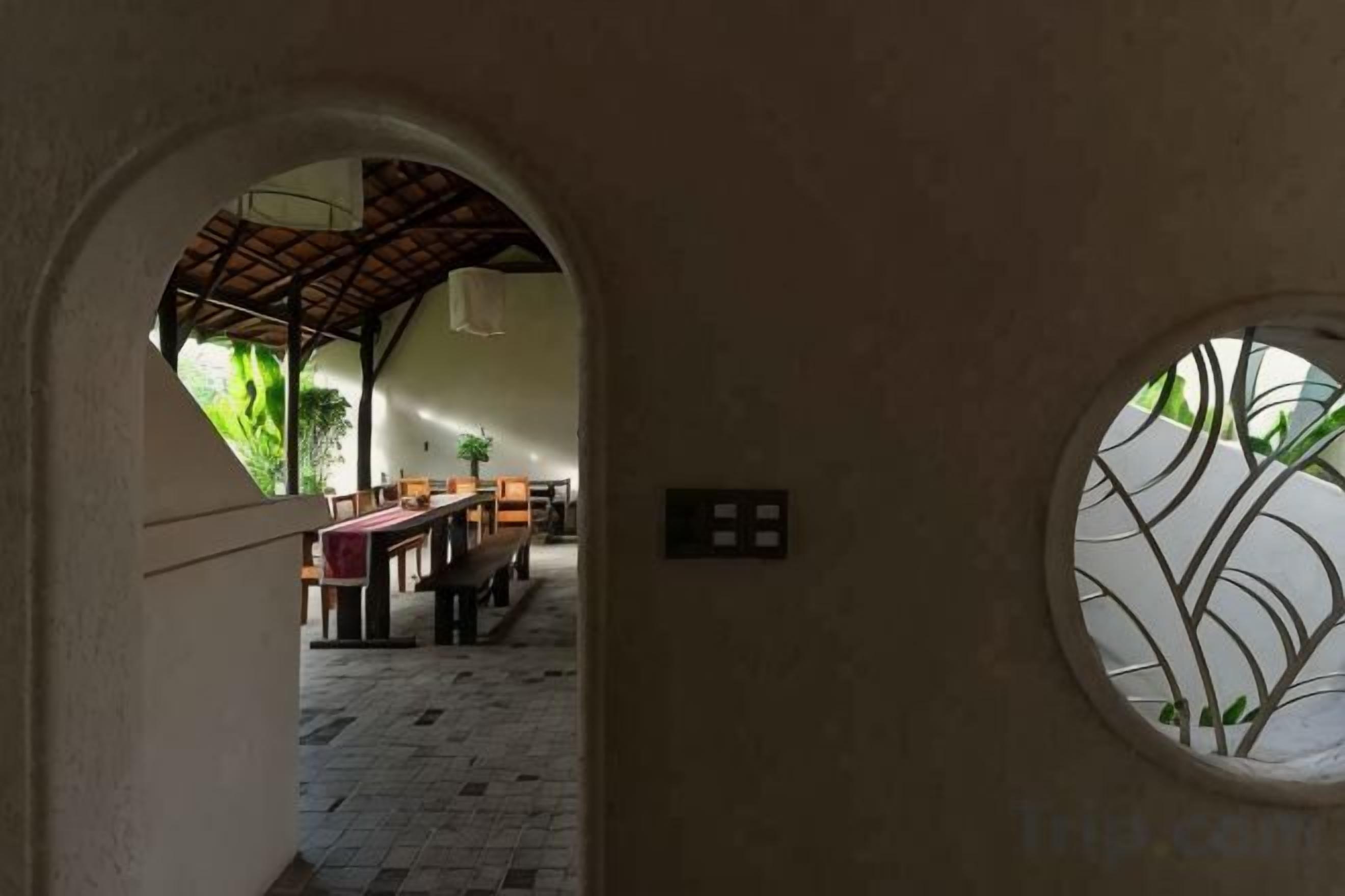 Vista interior de un edificio, con un área de comedor visible a través de un arco, una ventana redonda con un diseño decorativo y un techo con estructura de vigas de madera.