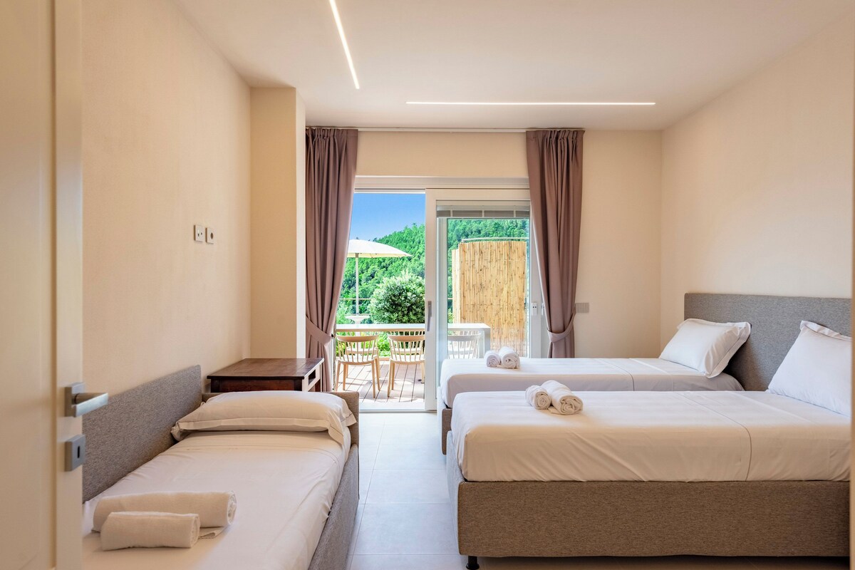 A hotel room with two beds, a balcony with a table and chairs, and a view of greenery.