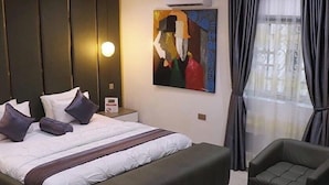 A modern hotel room with a large bed, a side table, a painting, a sofa, and a small round table. - The Oak Residence (Abuja)