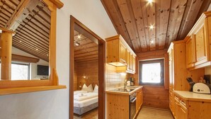 Un intérieur en bois comprenant un lit, un coin cuisine et une vue sur l’extérieur par une porte ouverte.