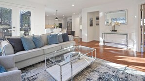 A modern living room with a glass coffee table, a sofa with blue cushions, and a view of the kitchen in the background. - Quiet 5BR Bend Retreat - Minutes to Downtown, Trails & More (Bend)