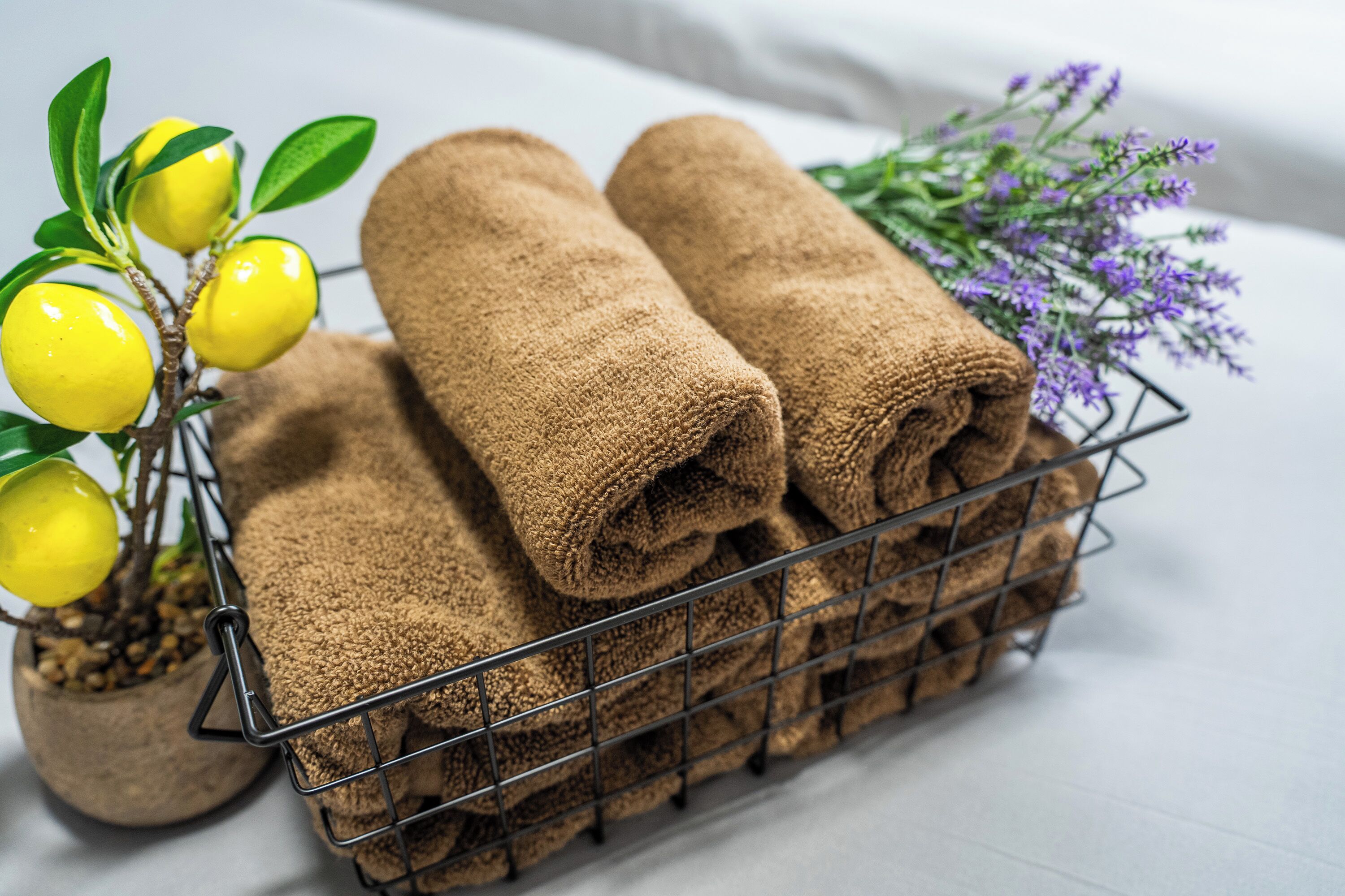 A metal basket containing rolled brown towels, a potted plant with yellow fruits, and purple flowers.