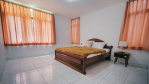 A bedroom with a wooden bed, two windows with orange curtains, a bedside table with a lamp, and a tiled floor.