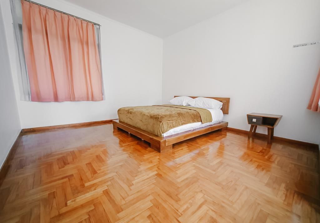 Quarto com cama de madeira, uma pequena mesa de cabeceira e janela com cortinas.