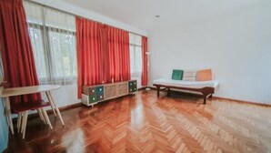 A room with a wooden parquet floor, a bed with a white mattress and colorful pillows, a small table with chairs, and a radiator.