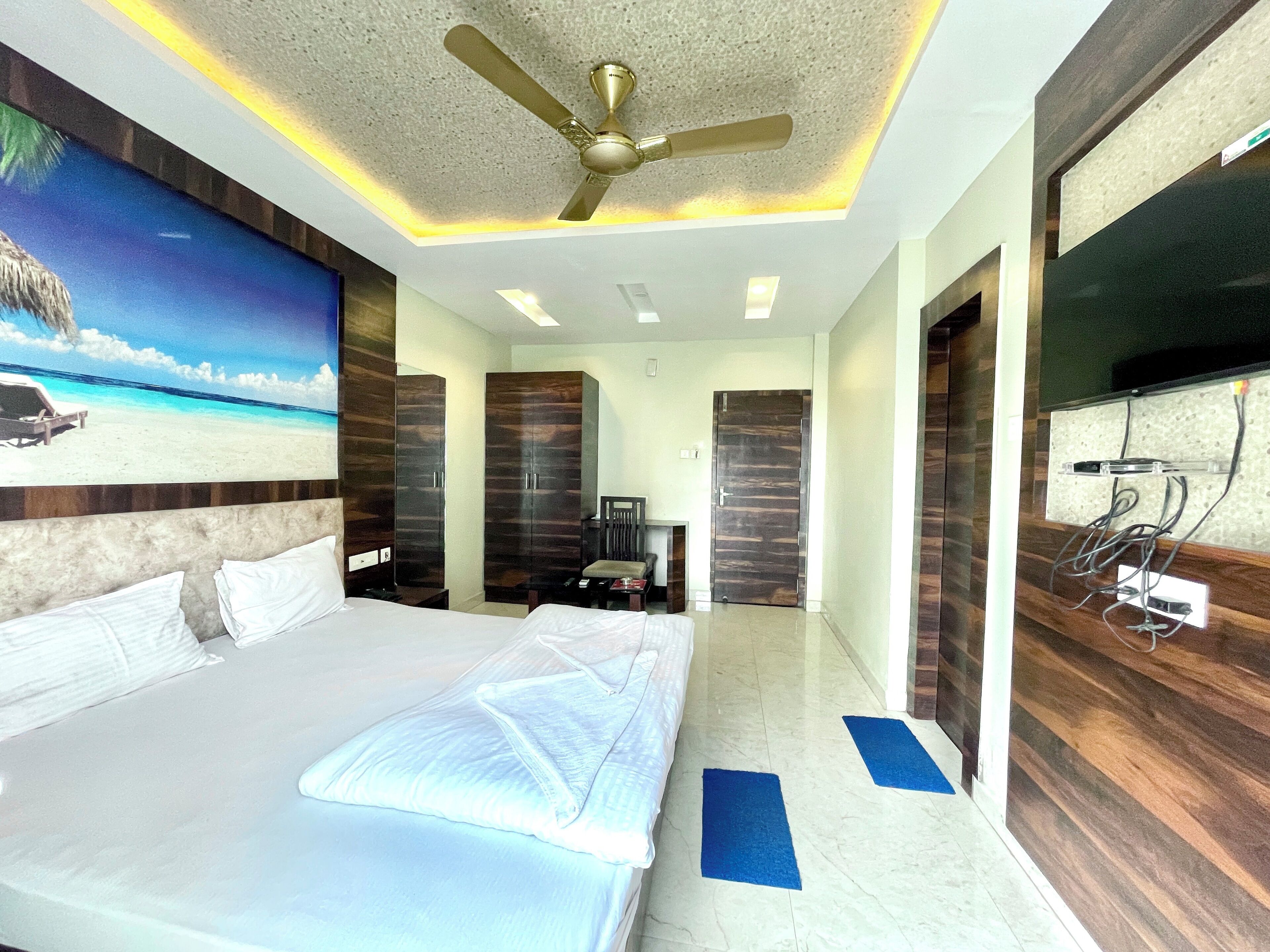 Una habitación de hotel con una cama, un ventilador de techo, un televisor montado en la pared y un cuadro con una escena de playa.