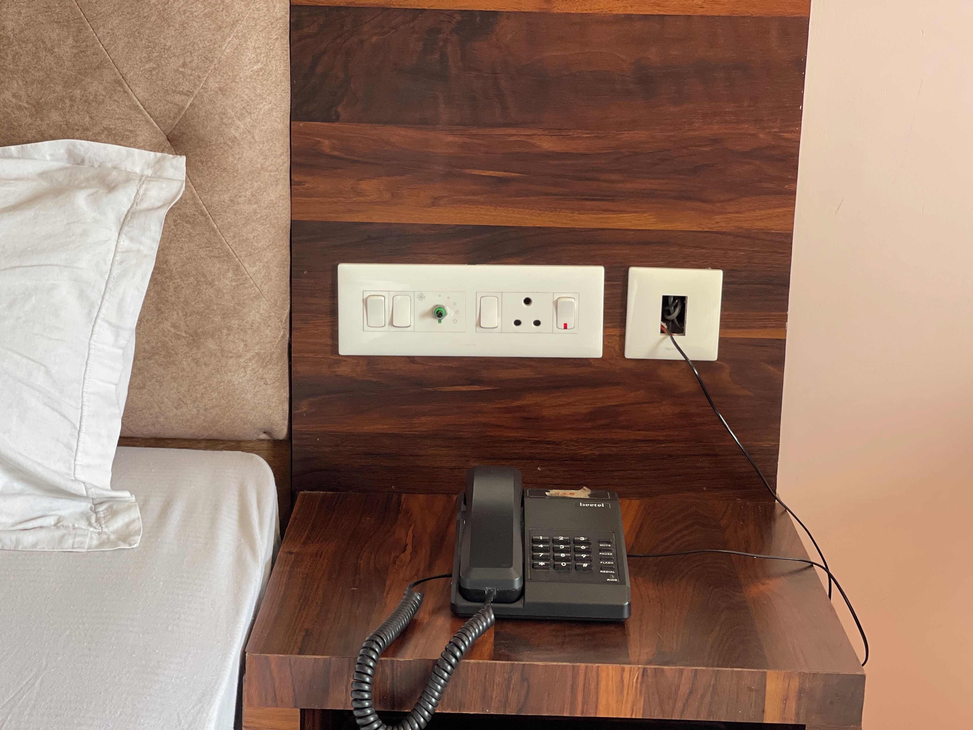 Habitación de hotel con cabecera de madera, cama con ropa de cama blanca, mesita de noche con teléfono e interruptores eléctricos.