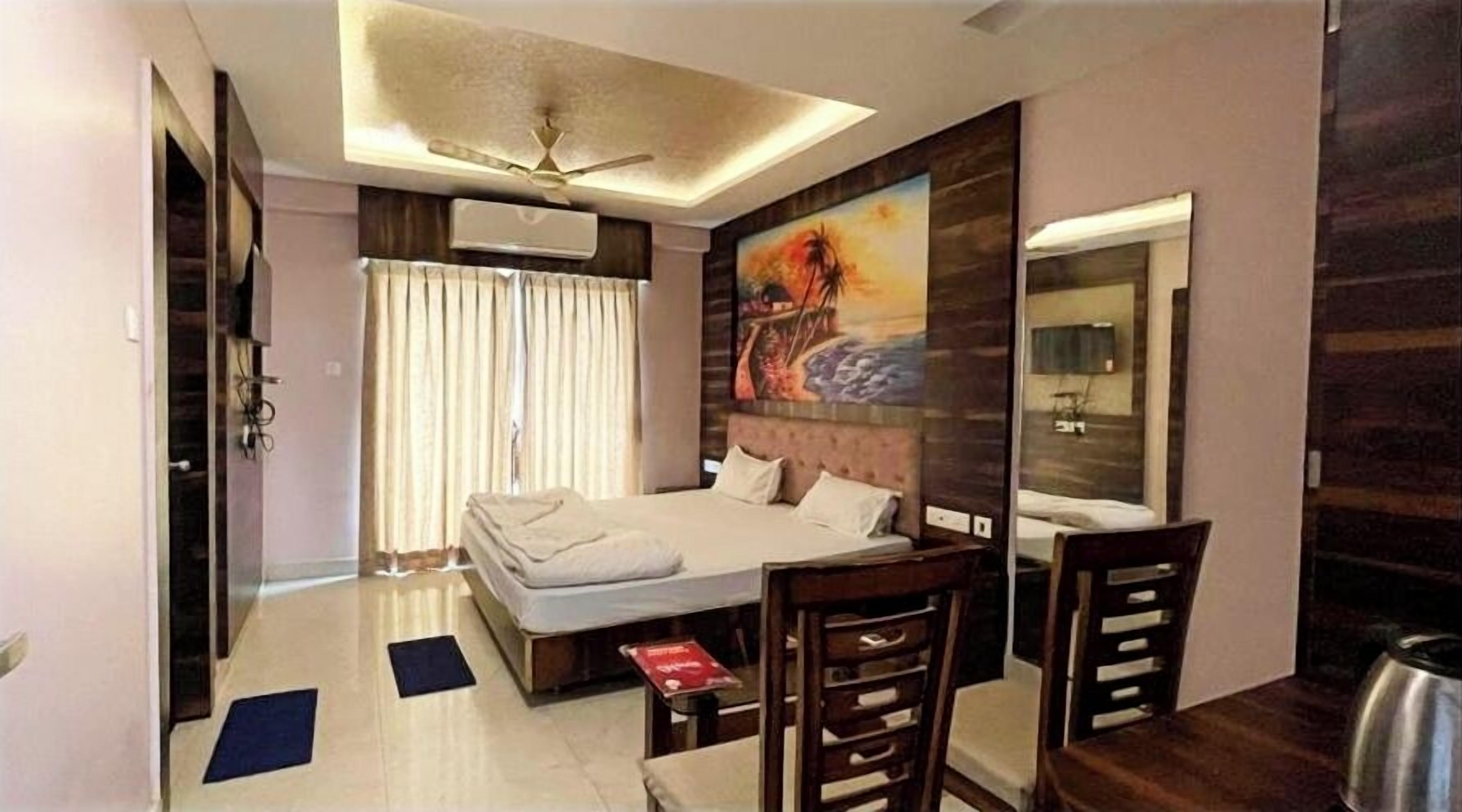 A hotel room with a bed, a desk, a chair, a ceiling fan, and a large painting on the wall.