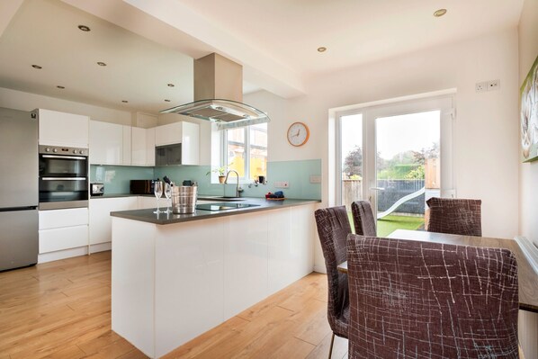 A modern kitchen with white cabinetry, a central island, and a dining area with brown upholstered chairs. - Elizabeth House (Kington)