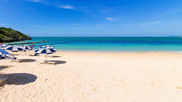Ein Strand mit klarem, blauem Wasser, weißem Sand und blauen Sonnenschirmen.