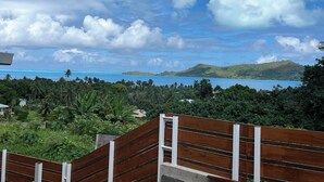 A view of a tropical landscape with a wooden fence, lush greenery, and a distant view of a blue sea and green hills. - A modern, well-equipped bungalow with an exceptional view of the lagoon! (Bora-Bora)