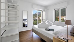 A modern bedroom with a large bed, a bedside table with a lamp, a wardrobe, and a view of the city through a large window. - Ulivo and Magnolia Bike & River Apartment (Arco)