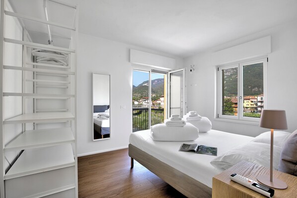 Ein modernes Schlafzimmer mit Bett, Nachttisch, Kleiderschrank und einem großen Fenster mit Blick auf Gebäude und Berge.