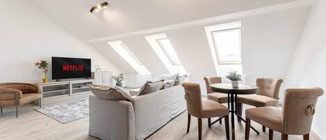 A modern loft apartment with a large sofa, a dining table, and a TV displaying 'NETFLIX'.