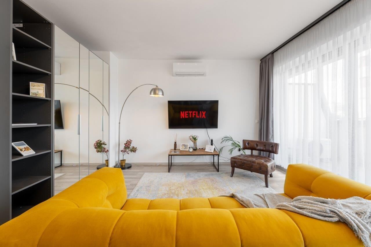Een moderne woonkamer met een gele bank, een houten salontafel en een flatscreen televisie aan de muur.
