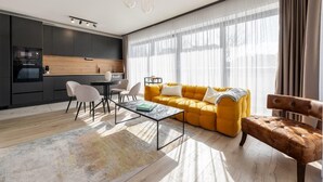 Una sala de estar moderna con sofá, mesa de centro y sillones. Se observa una zona de cocina con armarios y electrodomésticos.