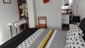 Un dormitorio con cama, estantería, silla, baño con lavamanos y espejo, y un cuadro en la pared.