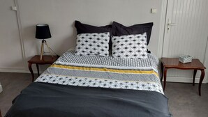 Un dormitorio con cama, mesita de noche con lámpara, una pequeña mesa con un objeto decorativo y una puerta.