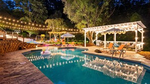 An outdoor swimming pool with a wooden deck, lounge chairs, and a pergola with string lights.