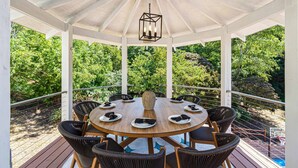 A covered outdoor dining area with a round table and chairs, surrounded by white pillars and a glass ceiling.