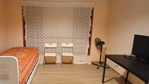 Ein kleines Zimmer mit einem Bett, zwei Stühlen, einem Schreibtisch, einem Fernseher und einem Ventilator.