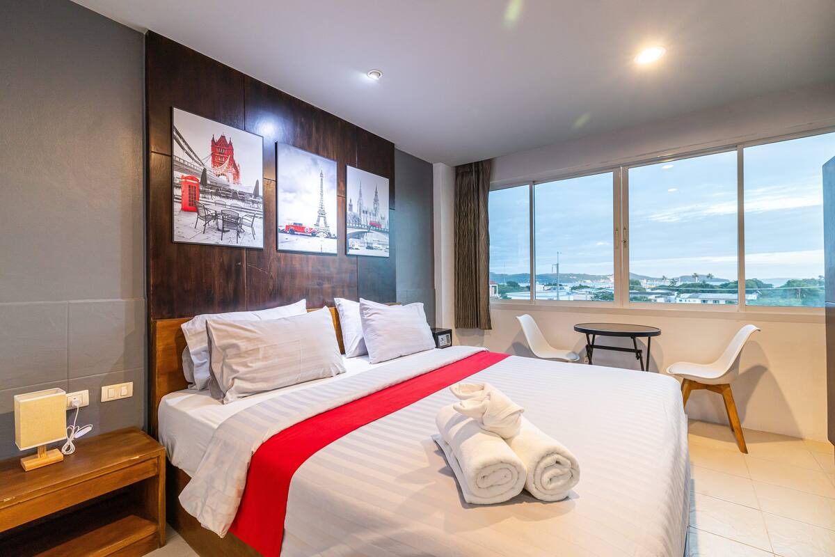 Quarto de hotel com uma cama grande, vista para a cidade, escrivaninha com abajur e uma cadeira.