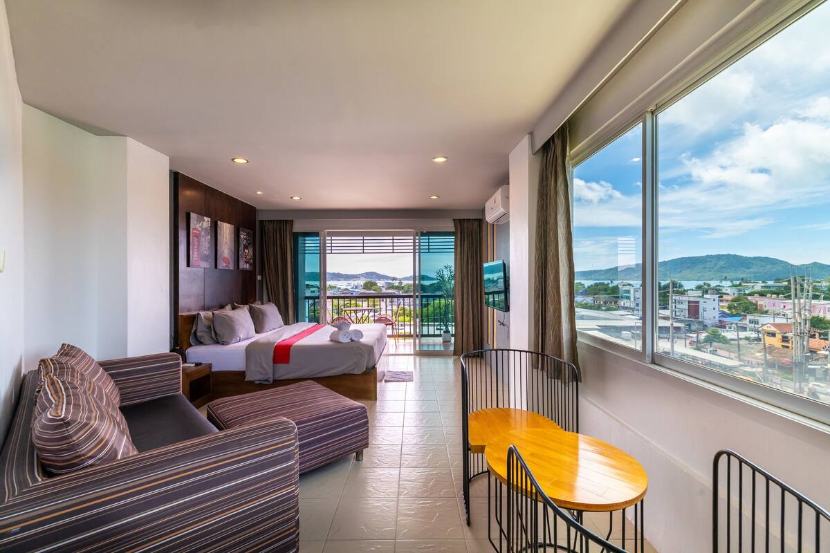 Quarto de hotel moderno com cama, sofá e mesa de jantar. Possui janelas amplas com vista para a cidade e varanda.