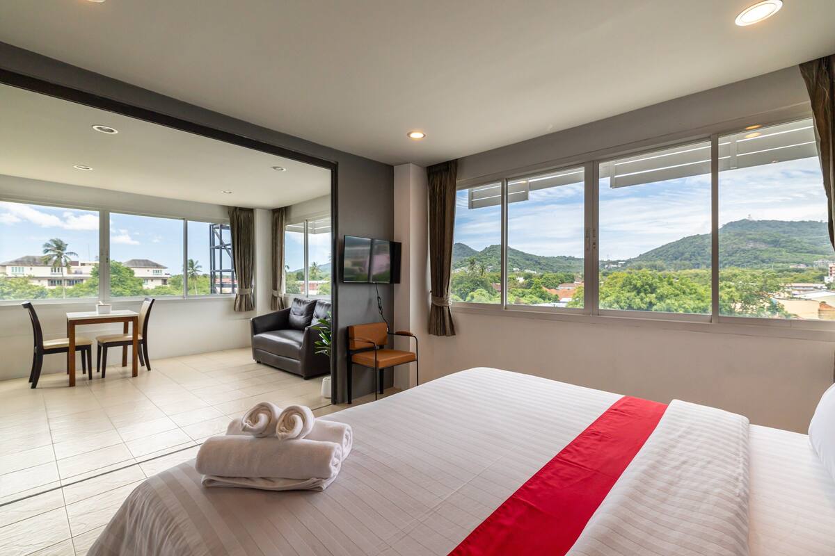 Quarto de hotel com uma cama grande, televisão, escrivaninha com cadeiras e vista para as montanhas.