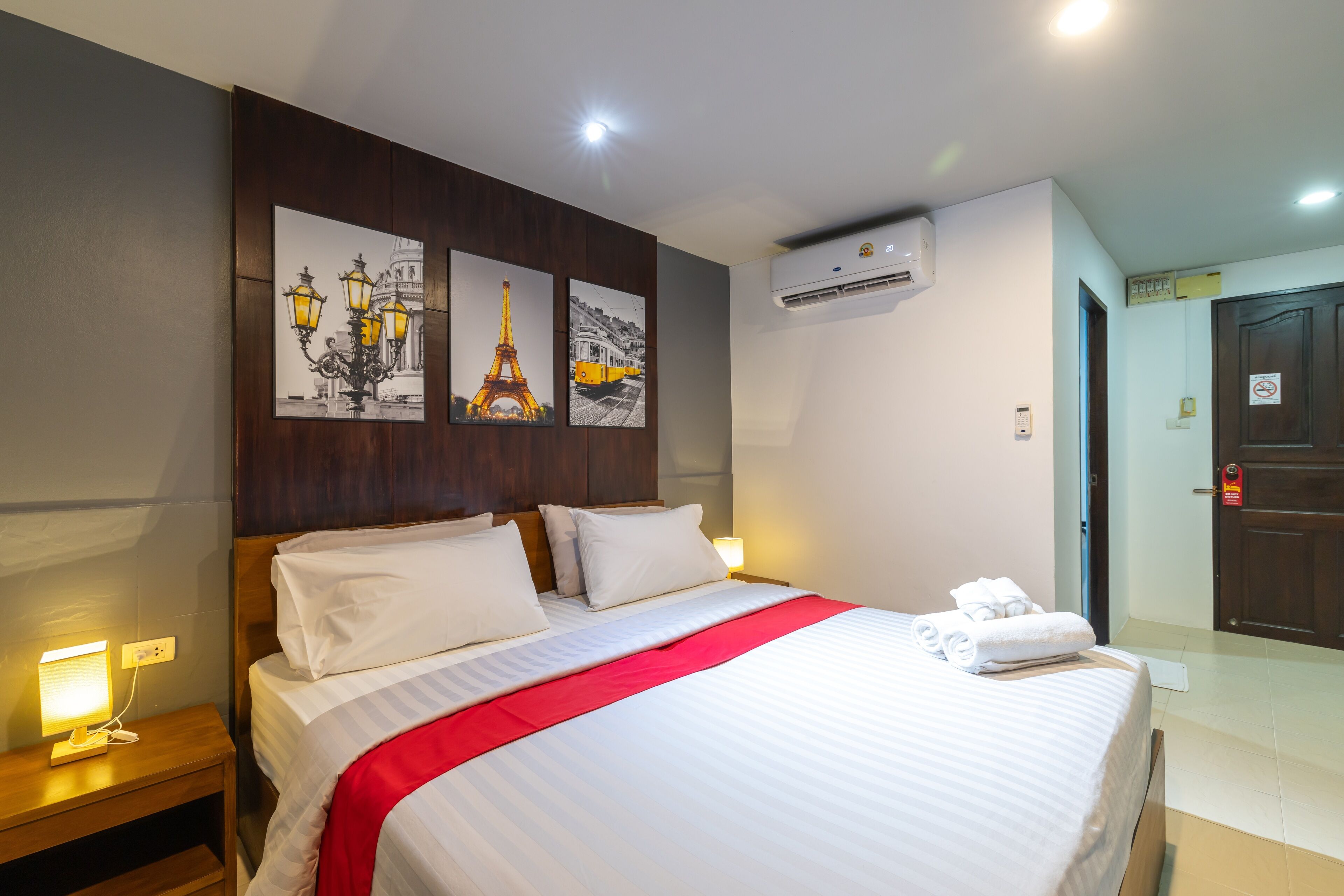 A neatly made bed with white linens and a red accent, a nightstand with a lamp, and three framed pictures on the wall depicting Paris landmarks.