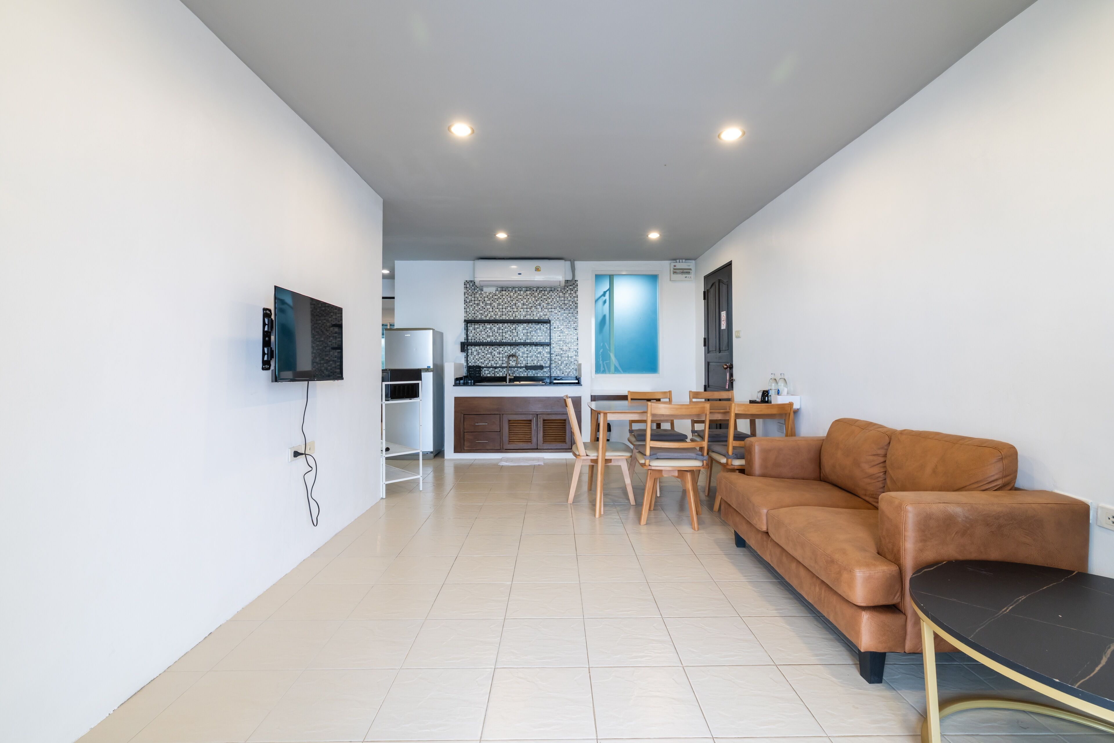 A modern living room with a brown leather sofa, a dining table with chairs, a flat-screen TV mounted on the wall, and a kitchen area in the background.