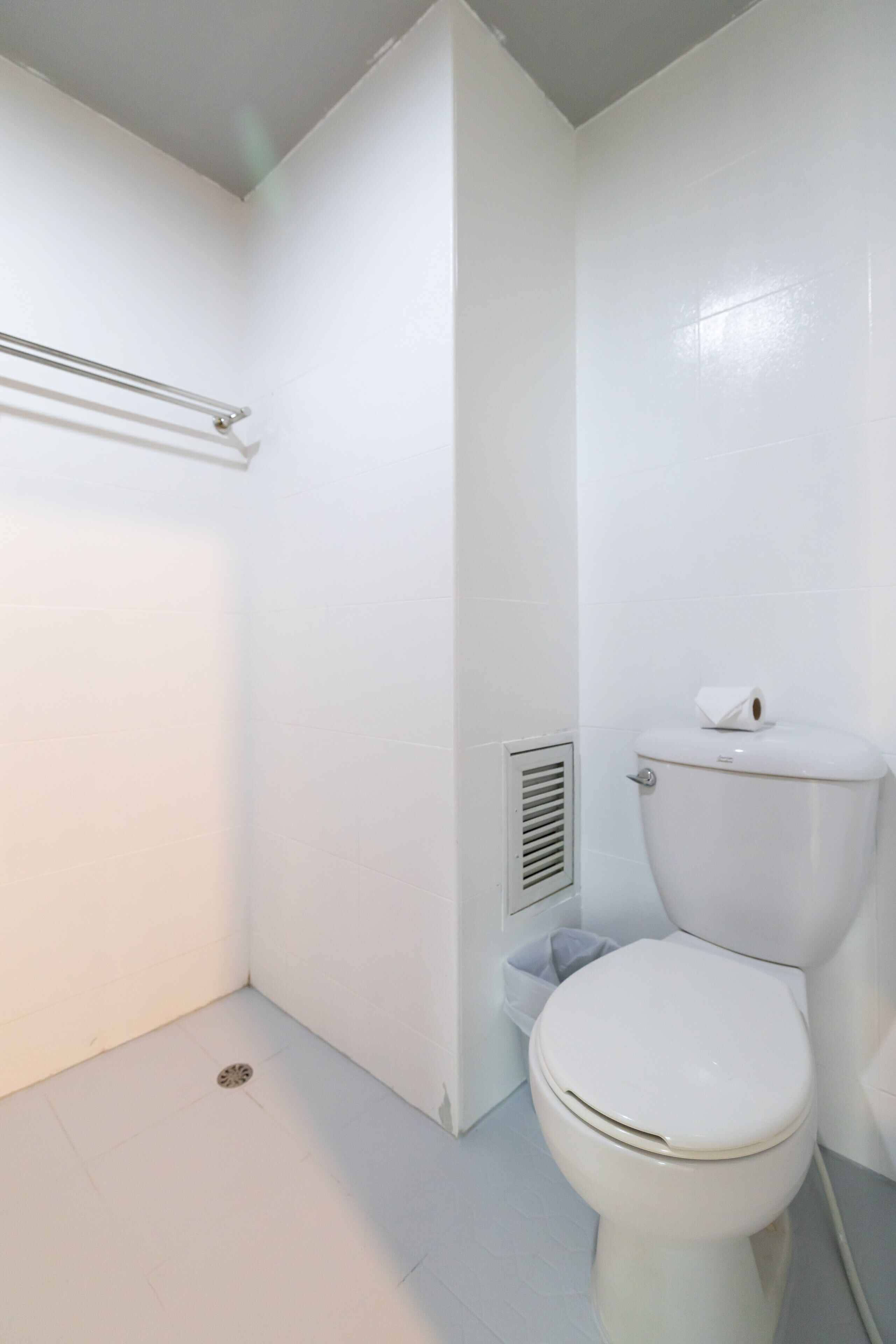 A small, white bathroom with a toilet, a shower area, and a vent on the wall.