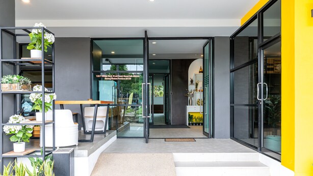 A modern building entrance with glass doors, a potted plant shelf, and a visible address sign. - Phuket Five Fortune Holiday Hotel (Rawai)