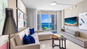 A modern living room with a sofa, a coffee table, and a TV. There is a large window with a view of a beach and buildings. - Luxury 2BR Apt With Ocean View, Pool & Amenities (Hollywood)
