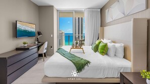A modern hotel room with a large bed, a desk, a TV, and a balcony with a view of the ocean and buildings. - Luxury 2BR Apt With Ocean View, Pool & Amenities (Hollywood)