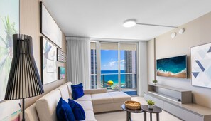 A modern living room with a sofa, a coffee table, and a TV. There is a large window with a view of a beach and buildings.