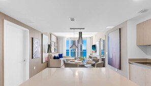 A modern hotel room with a large dining table, a kitchen area, and a view of the city skyline.