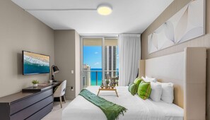 A modern hotel room with a large bed, a desk, a TV, and a balcony with a view of the ocean and buildings.